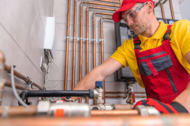 Caucasian Residential Heating Systems Technician in His 40s at Work. Hot and Cold Water Copper Made Pipelines in a Background.